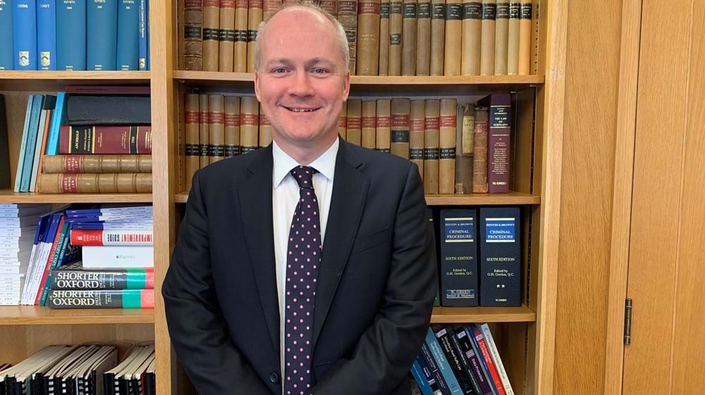 Image of Crown Agent John Logue who is smiling, standing in front of a wooden bookshelf